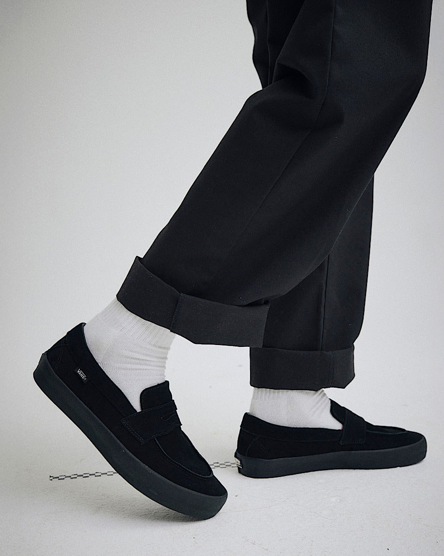 Person wearing black slip-on shoes, white socks, and black pants on a light gray background