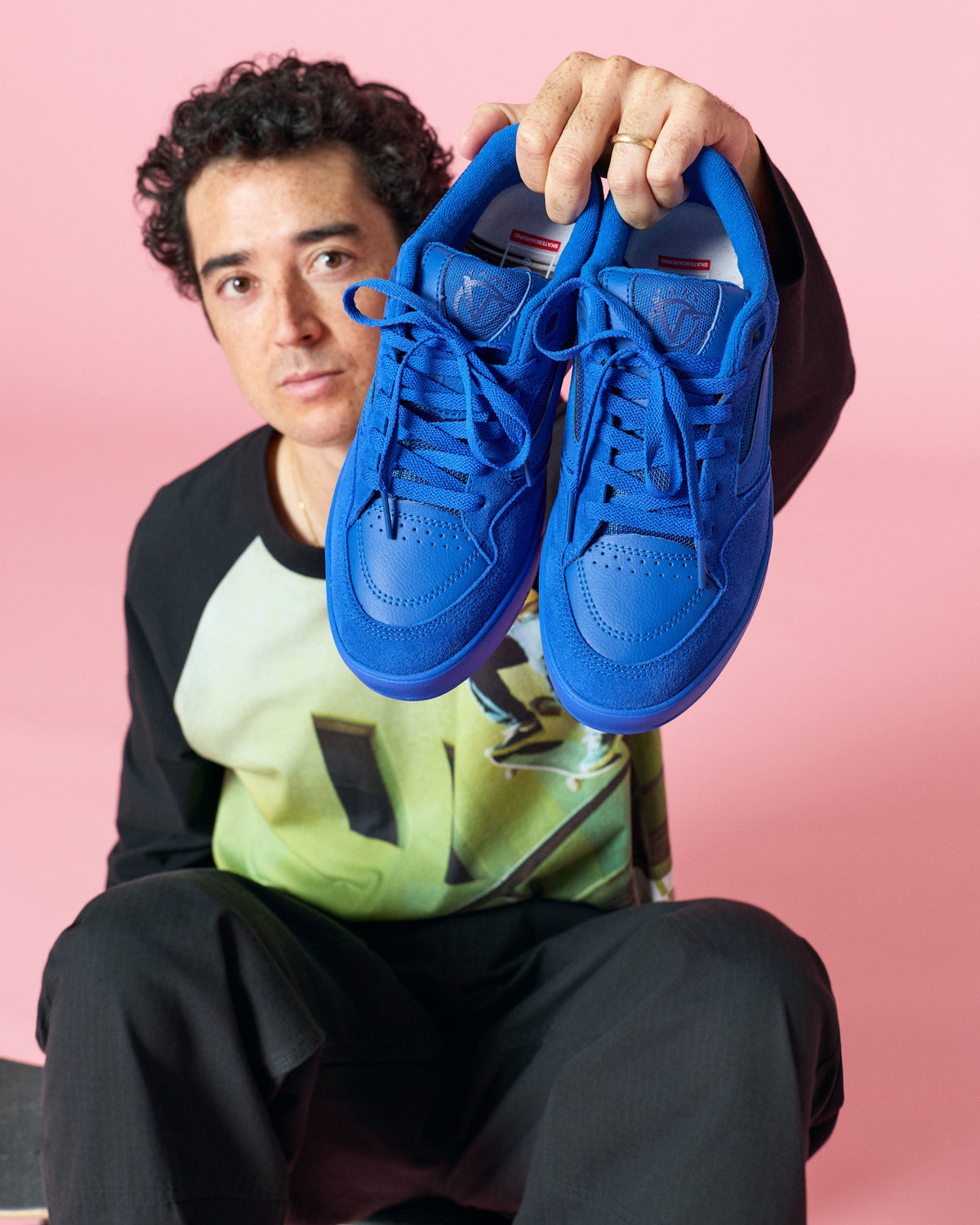 A man holding up a pair of bright royal blue sneakers against a solid pink studio background.