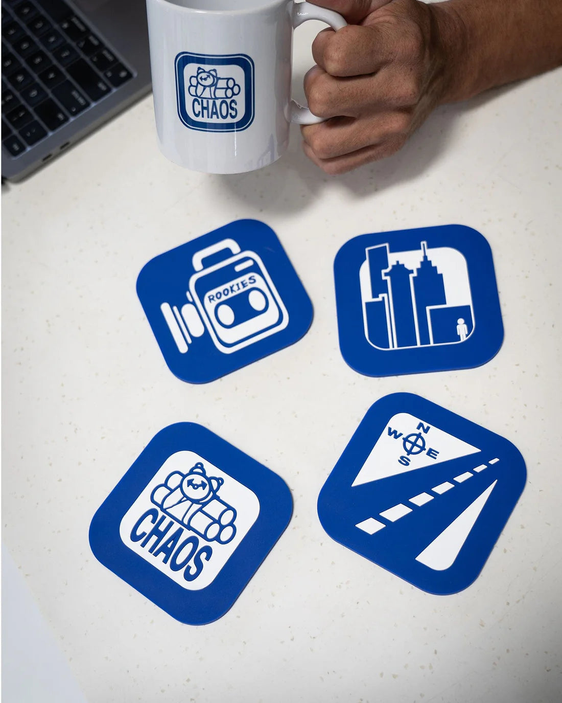 Four blue coasters with white designs on a white surface, one of which is held by a hand with a mug.