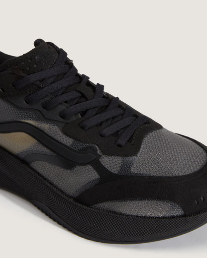 A close-up of a black and dark gray sneaker with textured mesh panels, black laces, and a chunky black sole, photographed on a light beige background.