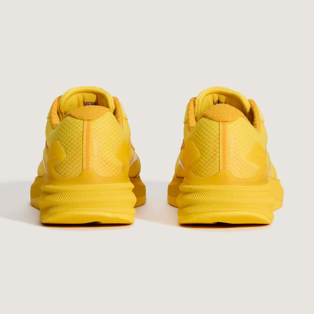 A pair of bright yellow athletic sneakers shown from the back, featuring textured mesh panels and chunky sole designs, set against a light gray studio background.