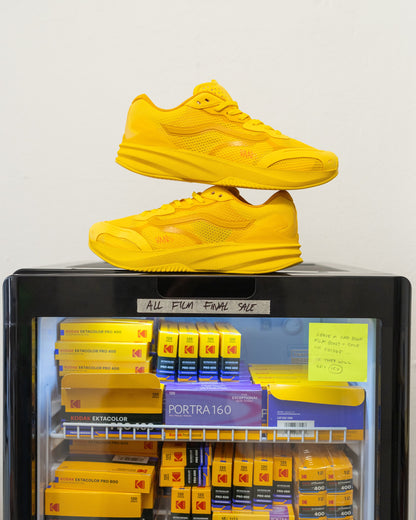 A pair of bright yellow athletic sneakers stacked on top of a black glass-front display case filled with Kodak Portra 160 film boxes and sale signs.