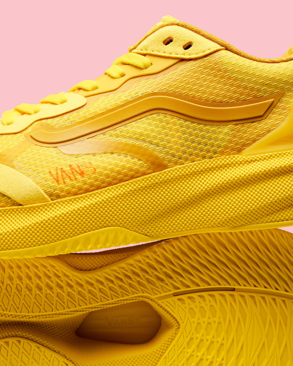 A close-up studio shot of a yellow Vans sneaker stacked on top of another shoe, with a solid pink background. The Vans logo is visible in orange on the side.