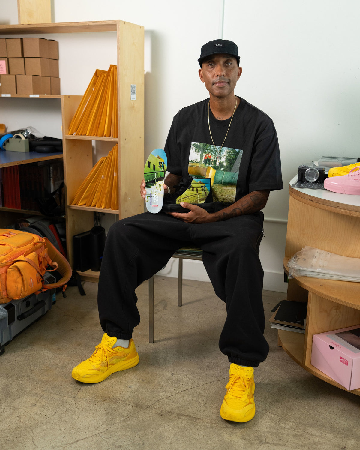 A man sits on a chair in a room, wearing a black graphic t-shirt, black sweatpants, and bright yellow sneakers, while holding a skateboard with a colorful graphic deck.