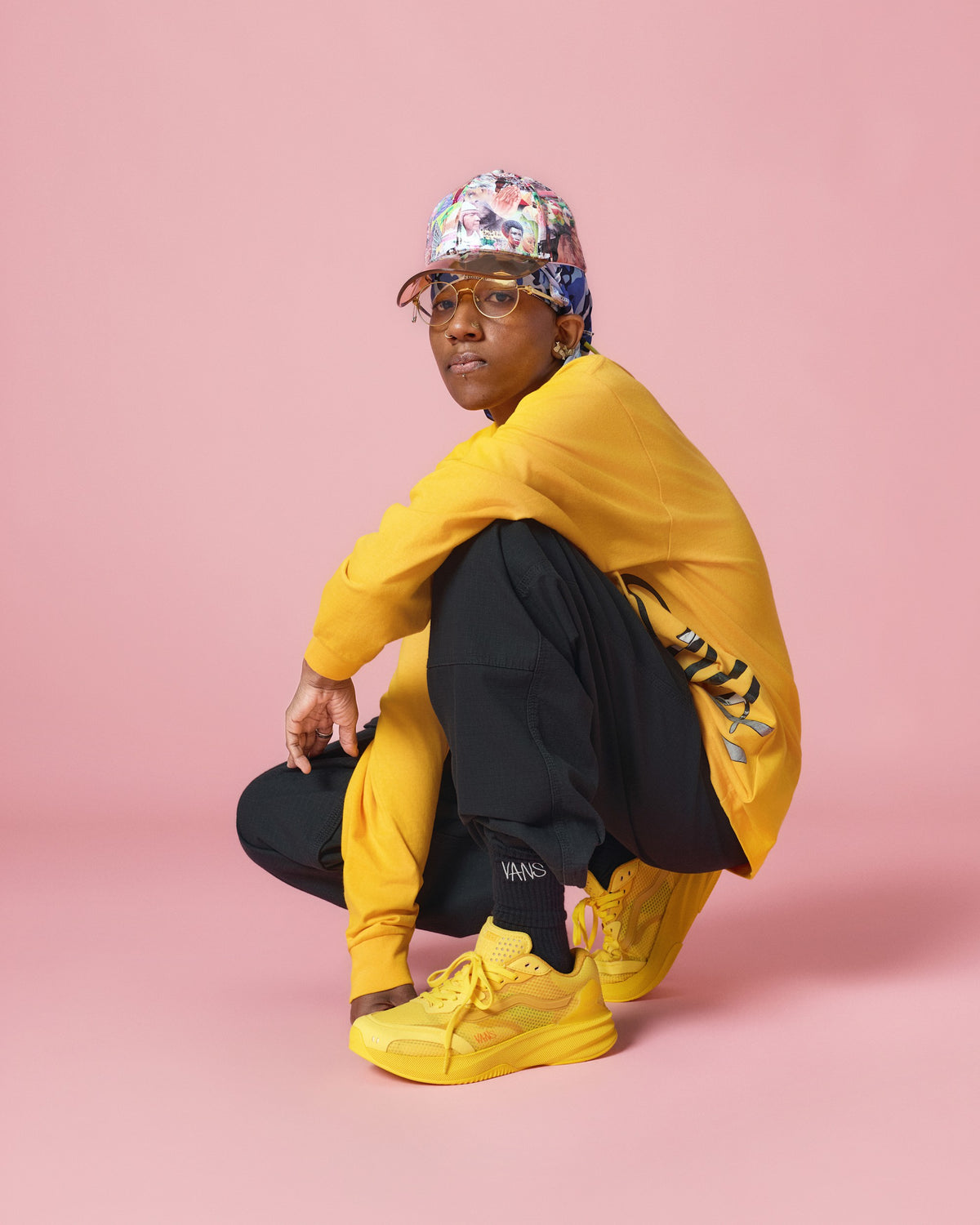 A model crouches in a studio setting against a solid pink background, wearing a bright yellow oversized sweatshirt, matching yellow joggers and sneakers, black sweatpants, and a colorful patterned cap.