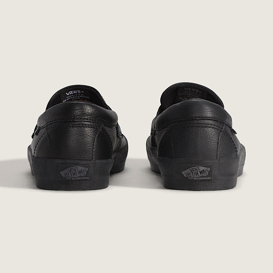 Black leather Vans slip-on sneakers shown from the back, featuring the classic Vans logo on the heel and tongue, set against a plain light beige background.