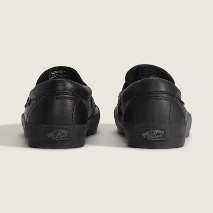 Black leather Vans slip-on sneakers shown from the back, featuring the classic Vans logo on the heel and tongue, set against a plain light beige background.