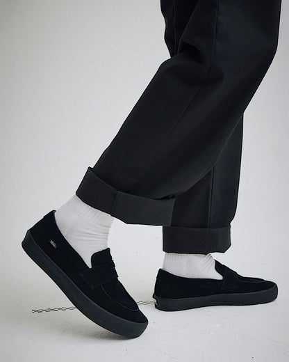 Person wearing black slip-on shoes, white socks, and black pants on a light gray background