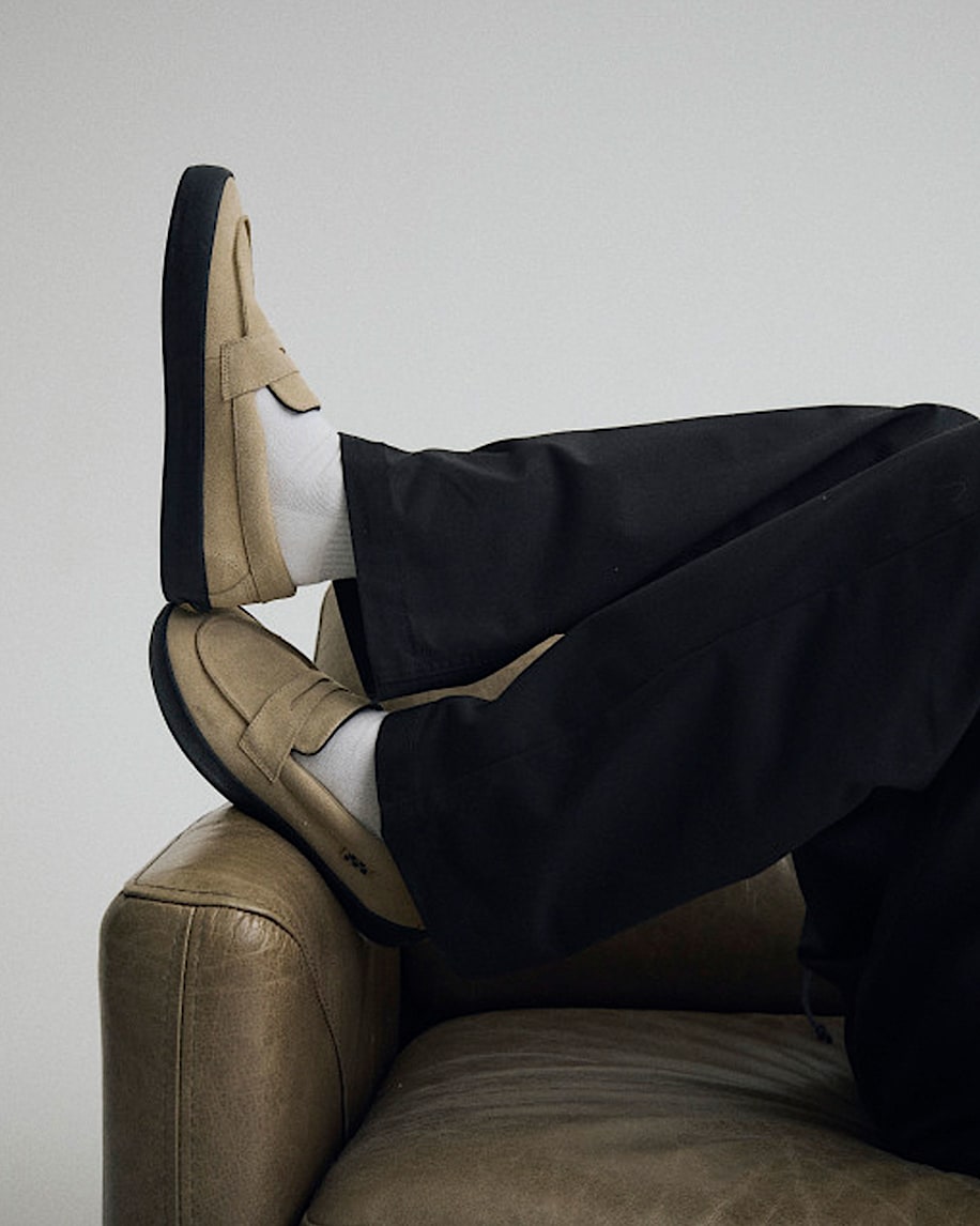 Person wearing black pants and tan loafers sitting on a brown leather couch.