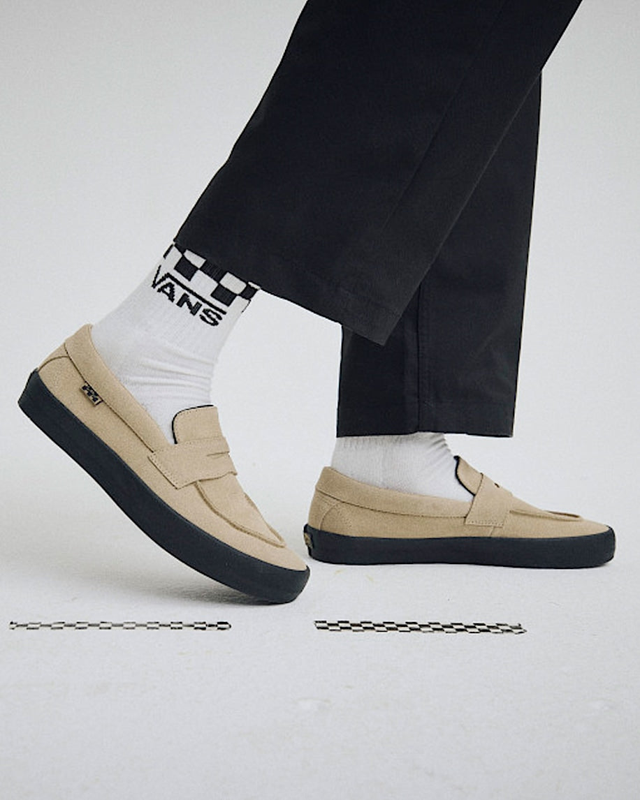 Beige loafers worn with black pants and white socks featuring a checkered pattern and brand logo.