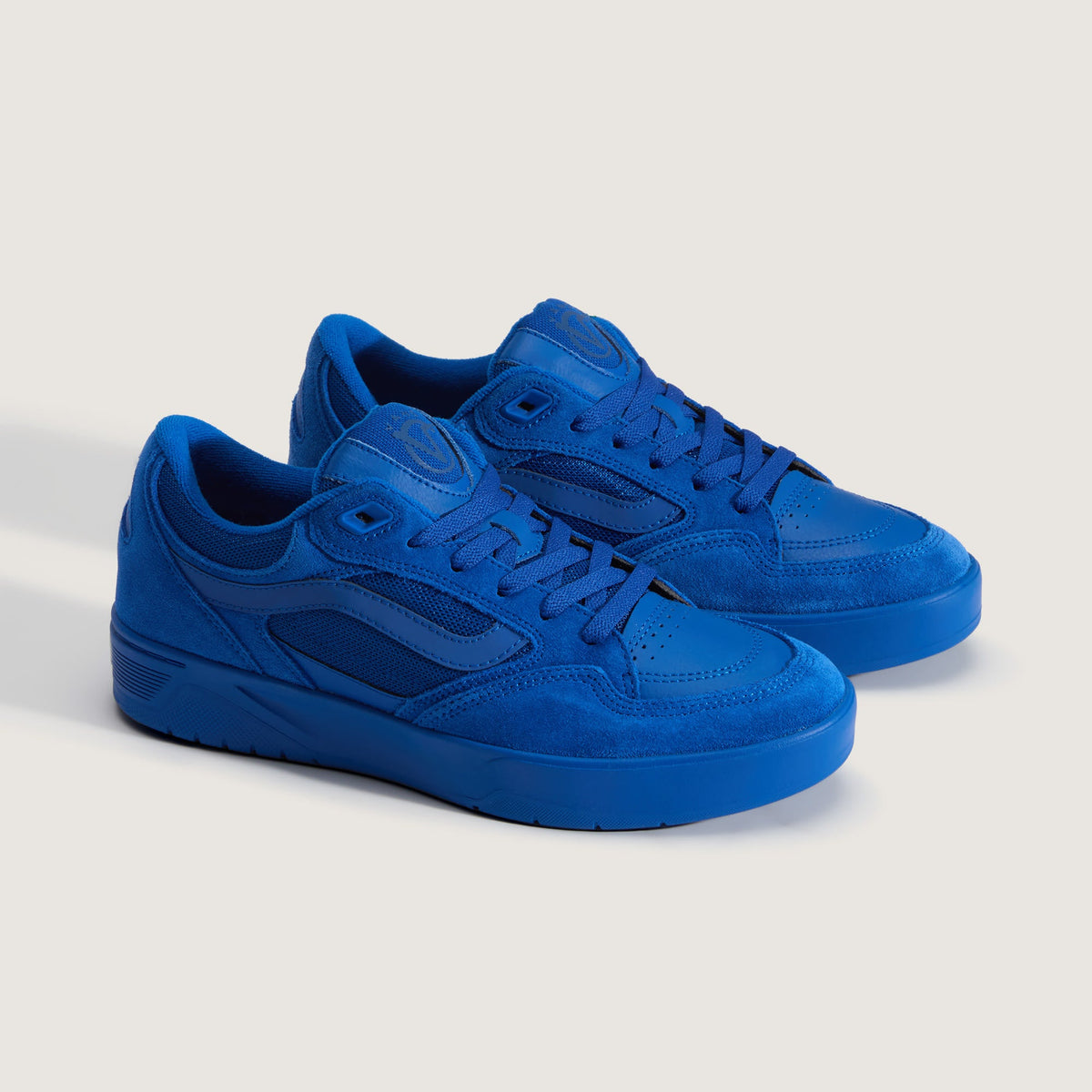 A pair of low-top sneakers in a monochromatic royal blue colorway, featuring suede and leather panels, shown on a light beige studio background.