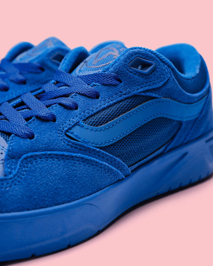 A close-up shot of a blue suede and mesh sneaker with matching blue laces and sole, set against a solid pink background.