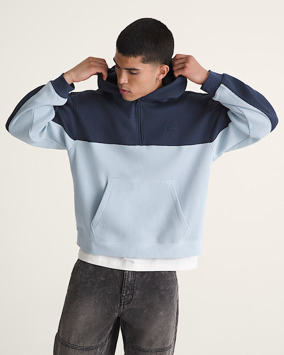 A man wearing a color-blocked half-zip hoodie with a light blue body, navy blue shoulders and hood, and dark gray jeans, posing against a light beige studio background.