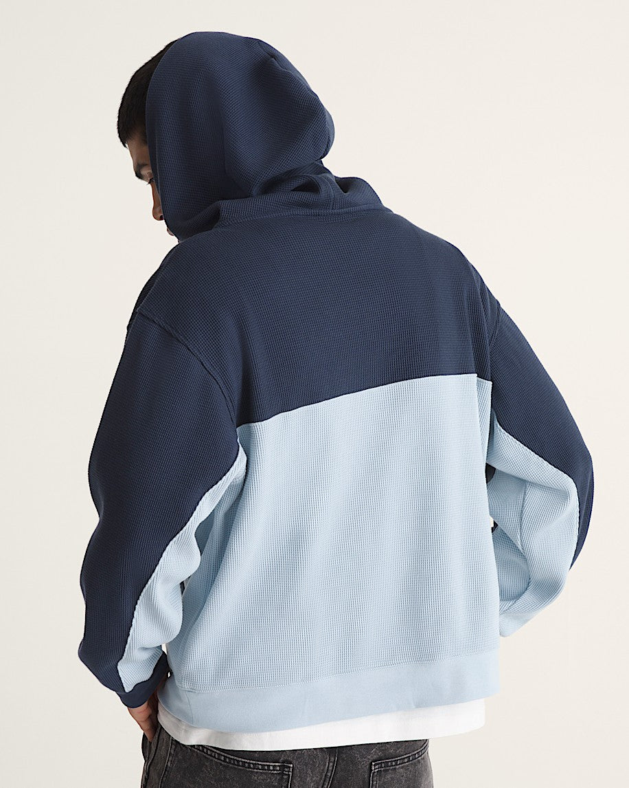 A person shown from the back wearing a colorblock hoodie with a navy blue top half and a light blue bottom half, paired with dark jeans against a plain off-white background.