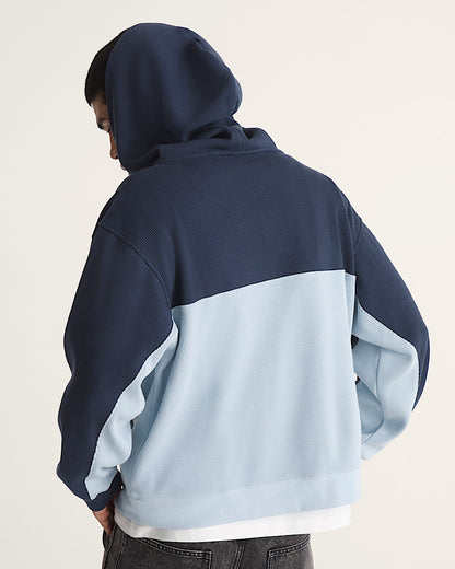 A person shown from the back wearing a colorblock hoodie with a navy blue top half and a light blue bottom half, paired with dark jeans against a plain off-white background.