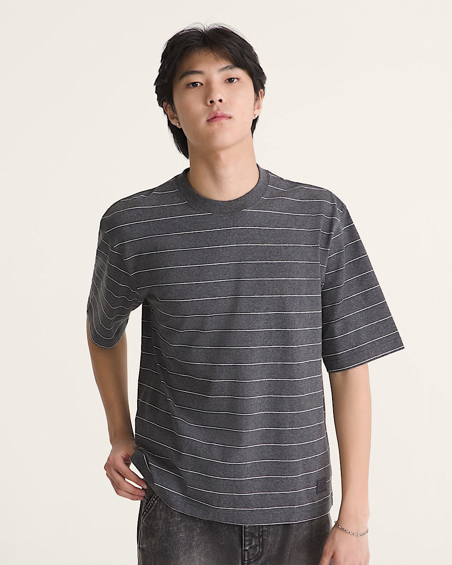A man wearing a dark gray crewneck t-shirt with thin white horizontal stripes, paired with dark gray jeans, photographed against a solid off-white studio background.