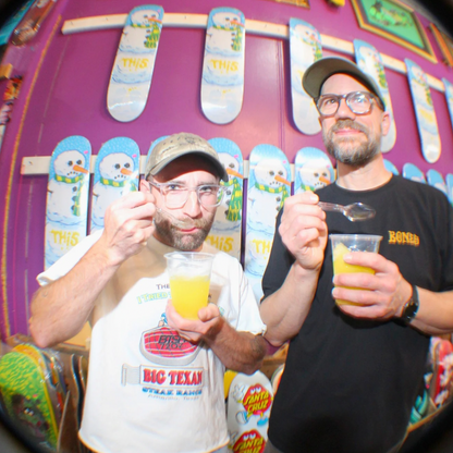 Two men smiling and holding up clear cups filled with orange-yellow drink, viewed through a circular black frame. One man wears a white t-shirt with a red and blue 'BIG TEXAS' graphic, and the other wears a black t-shirt with yellow text.